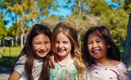 three smiling girls with long hair in the sunshine there is grass and trees behind them